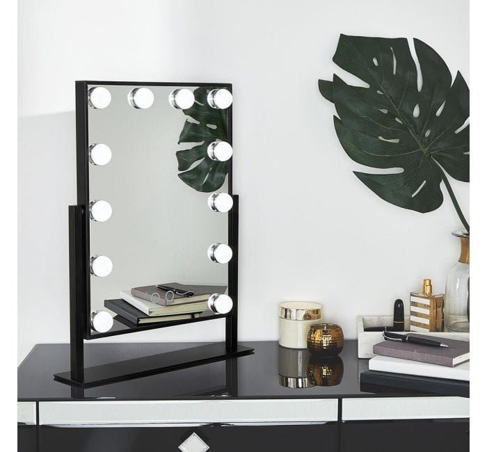 The UNIQ New York Mega makeup mirror with 12 bulbs by UNIQ sits on a black dresser, accompanied by books, a notebook, candles, and a large green monstera leaf in a vase.
