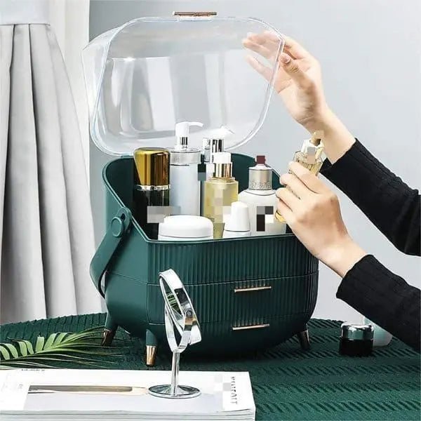 A person opens the UNIQ Retro Makeup Box Organiser in Dark Mint/Dark Green, revealing skincare and cosmetic bottles, jars, and pumps; a round mirror and brush from UNIQ sit on a green table nearby.