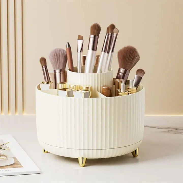 The UNIQ Rotating Makeup Organiser in Cream White by UNIQ features ribbed sides, gold legs, and multiple compartments for storing brushes, lipsticks, and pencils. It sits stylishly on a marble surface with a notepad beside it.