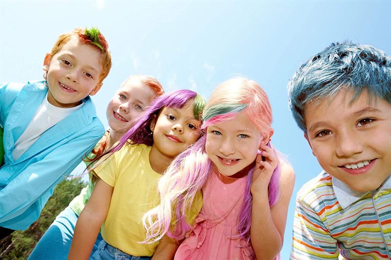 Five smiling children with vibrant blue, pink, green, purple, and red hair from the UNIQ Hair chalk - Vibrant Hair Chalk Color Pen set (12 colours) stand close together outdoors under a clear sky, looking down at the camera.