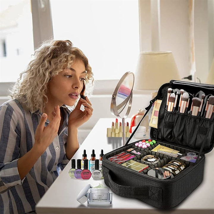 A woman with curly blonde hair applies makeup at a white vanity table filled with cosmetics. In the foreground is the UNIQ makeup travel bag—a black cosmetic bag by UNIQ with adjustable compartments holding brushes, palettes, and bottles.